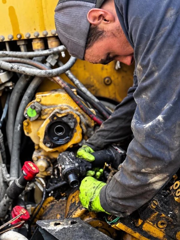 A mechanic in work gloves repairing hydraulic components on yellow heavy equipment